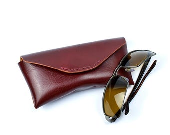May include: Sunglasses and a burgundy leather case. The sunglasses have a dark frame and brown-tinted lenses. The case is rectangular with a curved top and stitched edges. The items are set against a white background.