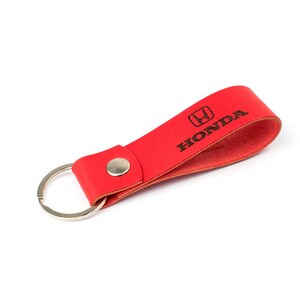 Könnte beinhalten: Roter Leder-Schlüsselanhänger mit silbernem Schlüsselring und Druckknopfverschluss. Der Schlüsselanhänger zeigt das Honda-Logo und das Wort "HONDA" in Schwarz.