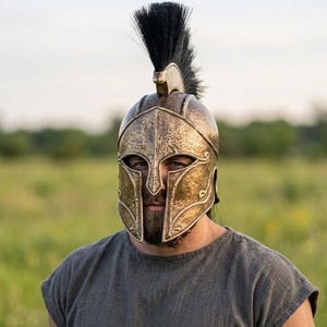 May include: A bronze-coloured Spartan helmet with detailed patterns and a black plume. The helmet has a face guard and a crest. The person is wearing a grey tunic. The background is a field of green grass.