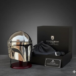 Op de afbeelding: Een gepolijste zilveren Mandalorian helm replica, gepresenteerd op een bordeauxrode standaard. De helm staat naast een zwarte doos met de tekst "MANDALORIAN FORGE". Een zwart trekkoordzakje en een riem zijn zichtbaar in de doos.