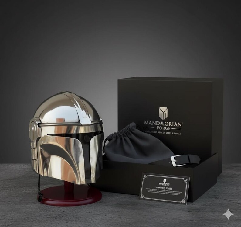 Op de afbeelding: Een gepolijste zilveren Mandalorian helm replica, gepresenteerd op een bordeauxrode standaard. De helm staat naast een zwarte doos met de tekst "MANDALORIAN FORGE". Een zwart trekkoordzakje en een riem zijn zichtbaar in de doos.