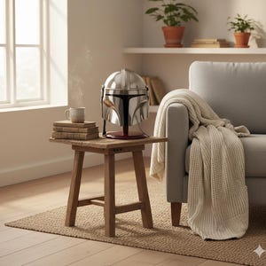 Op de afbeelding: Een zilveren Mandalorian helm replica op een rode standaard staat op een kleine houten tafel. Een stapel boeken en een mok staan ook op de tafel. De sc&egrave;ne speelt zich af in een lichte kamer met een raam en een bank.