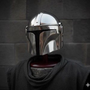 Op de afbeelding: Een glanzende, zilveren Mandalorian helm met een T-vormig vizier. De helm wordt getoond op een donkergrijze hoodie, tegen een donkergrijze bakstenen muur. Het gepolijste oppervlak van de helm reflecteert licht.