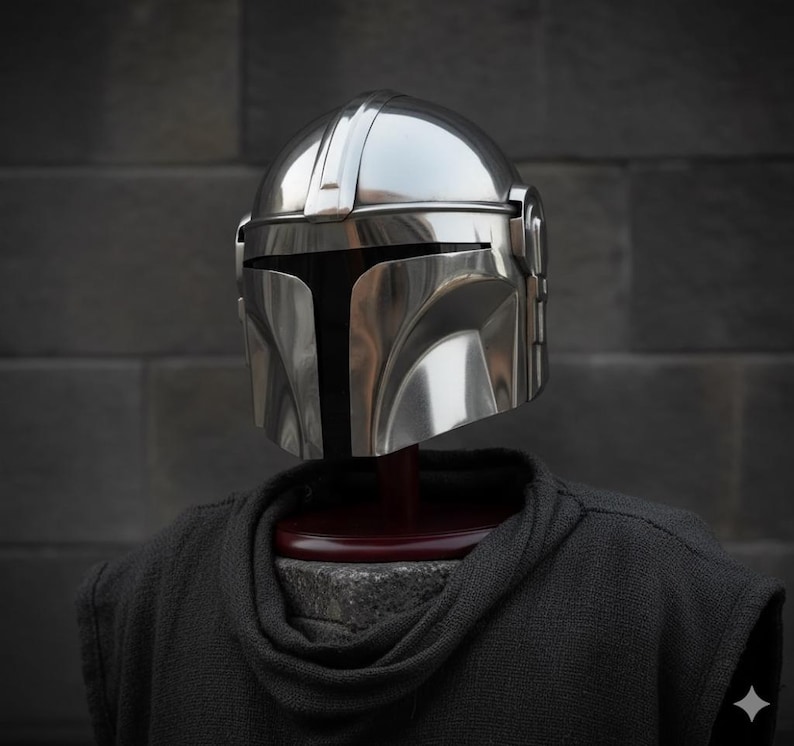 Op de afbeelding: Een glanzende, zilveren Mandalorian helm met een T-vormig vizier. De helm wordt getoond op een donkergrijze hoodie, tegen een donkergrijze bakstenen muur. Het gepolijste oppervlak van de helm reflecteert licht.