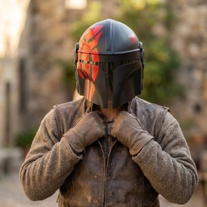 May include: A black and red helmet with a distinctive design, worn with a brown leather jacket and gloves. The helmet features red markings and a visor. The person is wearing a brown jacket and gloves.