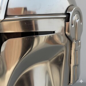 Op de afbeelding: Close-up van een metallic zilveren helm met een reflecterend oppervlak. De helm heeft een vizier en een gedetailleerd ontwerp, wat wijst op een futuristisch of sciencefiction thema. Het oppervlak van de helm vertoont lichte slijtage.