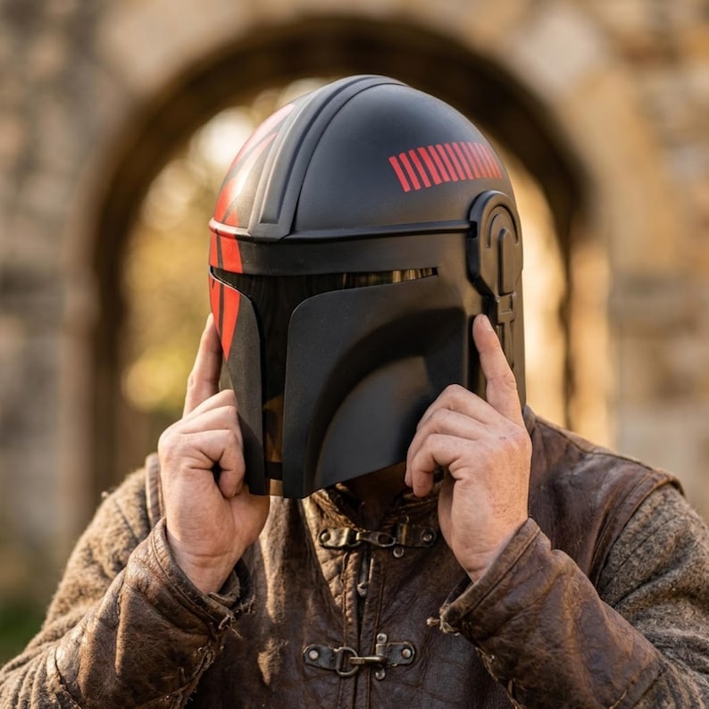 May include: Black helmet with red stripes, reminiscent of a Mandalorian design. The helmet features a visor and is being held by a person wearing a brown leather jacket. The background includes an arched stone structure.