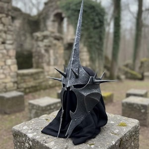 Puede incluir: Un casco de fantasía gris oscuro con una larga espiga central y múltiples espigas más pequeñas. El casco descansa sobre una superficie de piedra, con tela negra debajo. El fondo presenta una antigua estructura de piedra y árboles.