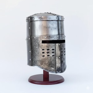 May include: A silver medieval helmet with a rectangular visor and ventilation slots. The helmet is riveted and mounted on a dark wooden stand, showcasing its historical design and craftsmanship.