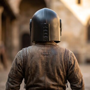 May include: A person wearing a black helmet and a brown leather jacket, viewed from the back. The helmet has a rounded top and rectangular details on the side and back. The jacket has a weathered appearance.
