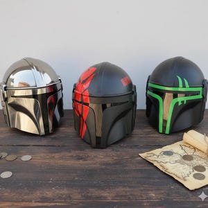 May include: Three Mandalorian helmets in a row. The first is chrome, the second is black with red accents, and the third is black with green accents. A rolled-up map and coins are on the wooden surface.