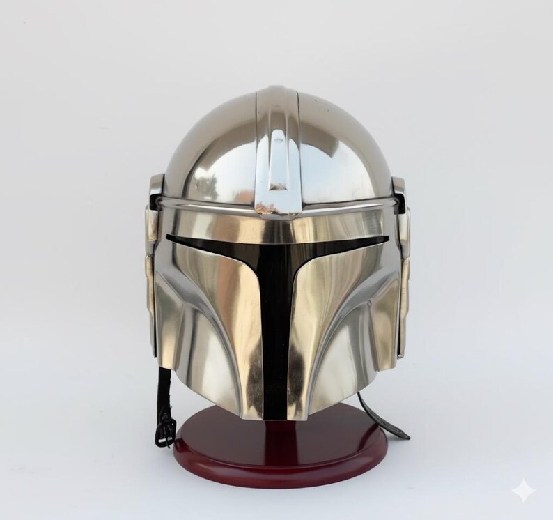 Op de afbeelding: Een metallic zilveren Mandalorian helm met een glad, reflecterend oppervlak. De helm heeft een T-vormig vizier en staat op een donkerrode houten standaard. Het ontwerp doet denken aan een populair sciencefictionpersonage.