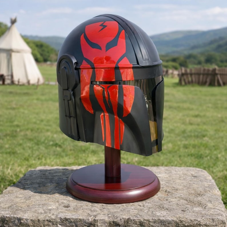 May include: A black helmet with red accents, displayed on a wooden stand. The helmet features a red design on the front and top, with a visor and side details. The stand is a dark brown color, and the background is a grassy field.