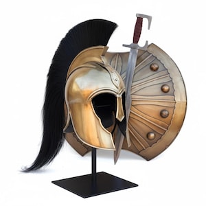 May include: A gold-colored Spartan helmet with a black plume, a matching shield, and a sword. The shield is bronze-colored with a sunburst design. The sword has a brown handle. The helmet is displayed on a black stand.