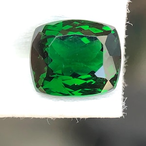 May include: A cushion-cut, faceted gemstone with a vibrant, deep green color. The gem is set against a white background, showcasing its clarity and brilliance. The gemstone's facets reflect light, creating a sparkling effect.