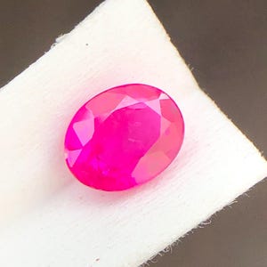Pode incluir: Uma gema rosa vibrante, facetada e de forma oval. A gema está sobre uma superfície branca e texturizada, com um fundo escuro. A lapidação da gema cria múltiplas superfícies refletoras, realçando o seu brilho. Para joalharia.