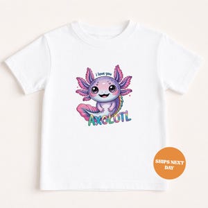 Könnte beinhalten: Weißes T-Shirt mit einer bunten Axolotl-Grafik. Der Aufdruck zeigt den Text "i love you" und "AXOLOTL" in bunten Buchstaben. Der Axolotl ist in Lila, Rosa und Blau dargestellt. Ein lustiges und lebendiges Design.