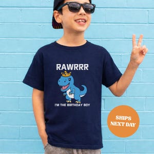 Op de afbeelding: Marineblauw T-shirt met een cartoon dinosaurus met een kroon. De tekst "RAWRRR" staat boven de dinosaurus en "I'M THE BIRTHDAY BOY" staat eronder. Het T-shirt wordt door een kind gedragen.