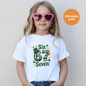 Op de afbeelding: Een wit T-shirt met een St. Patrick's Day-ontwerp met de groene cijfers "67", een kabouterhoed en de woorden "Six" en "Seven". Het T-shirt wordt gedragen door een jong persoon met een roze hartvormige zonnebril.