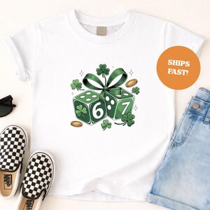 Op de afbeelding: Wit T-shirt met een groene afbeelding van twee dobbelstenen met de nummers 6 en 7, versierd met klavers en een groen lint. Gouden munten en klavers zijn verspreid rond de dobbelstenen. Het shirt wordt gecombineerd met een spijkerbroek en schoenen met ruitjes.