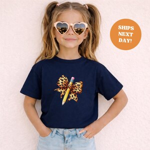 Op de afbeelding: Een marineblauw t-shirt met een potloodontwerp en een luipaardprint strik. De potlood is geel met een roze gum. Het meisje draagt hartvormige zonnebrillen.