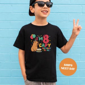 Op de afbeelding: Zwart T-shirt met een kleurrijk grafisch ontwerp voor een 8e verjaardag. Het ontwerp bevat de tekst "I'm 8 CAPY BIRTHDAY TO ME" met een cartoon capibara, ballonnen en een verjaardagstaart. Het shirt wordt door een kind gedragen.