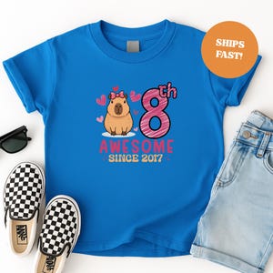 Op de afbeelding: Een blauw T-shirt met een cartoon capibara met een roze strik, het nummer 8 en de tekst "AWESOME SINCE 2017". De afbeelding bevat een spijkerbroekshort en zwart-wit geblokte instappers.