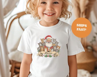 May include: White t-shirt featuring a Santa Claus graphic surrounded by cats. Santa wears a red hat and is surrounded by various colored cats. A banner below reads "Mason". An orange circle with the text "SHIPS FAST!" is visible.