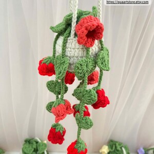 May include: A handmade crochet hanging planter in cream, adorned with red flowers and green leaves. The planter is suspended by a cream-colored rope. The text "QuaArtStore" and a website address are visible.