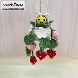 May include: A handmade crochet hanging planter featuring a smiling yellow bee with white wings, nestled in a beige pot. The pot is adorned with green leaves, white flowers, and red and pink crochet strawberries. The text "QuaArtStore" and the Etsy shop URL are visible.