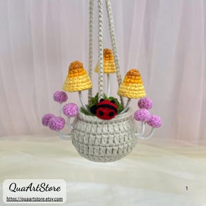 May include: A handmade crochet hanging planter featuring yellow and orange mushrooms, purple flowers, and a red ladybug. The planter is suspended by a cream-colored rope. The shop name is QuaArtStore.