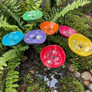 Puede incluir: Una colección de estacas de jardín con forma de flores de cerámica coloridas. Cada flor es de un color diferente, incluyendo verde, azul, morado, naranja, rosa, rojo y amarillo. Canicas de vidrio transparente están colocadas en el centro de cada flor.