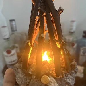 以下が含まれることがあります： ちらちらと光るミニチュアキャンプファイヤーの装飾。構造は、灰色の石の基盤の上に置かれた、中心の光源の周りに配置された濃い茶色の小枝で作られています。光の暖かい輝きは居心地の良い雰囲気を作り出します。