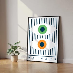 Könnte beinhalten: Gerahmter Bauhaus-Kunstdruck mit zwei stilisierten Augen mit grünen und orangefarbenen Pupillen auf gestreiftem Hintergrund. Das Wort "BAUHAUS" ist weiß am unteren Rand des schwarzen Rahmens gedruckt. Der Druck lehnt an einer weißen Wand.