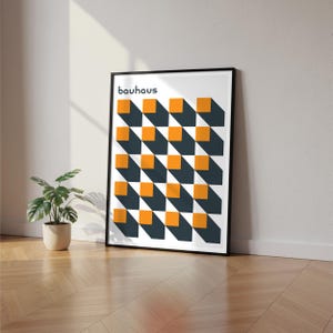 May include: Framed Bauhaus art print featuring a geometric pattern of orange squares and dark blue shadows. The word "bauhaus" is printed in black at the top. The artwork is displayed on a wooden floor next to a potted plant.