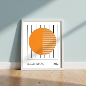 Op de afbeelding: Ingelijste Bauhaus-kunstprint met een geometrisch ontwerp in oranje en grijs. Het kunstwerk bevat een grote oranje cirkel met grijze horizontale lijnen en zwarte verticale lijnen. Het woord "BAUHAUS" en het jaar "1923" staan onderaan afgedrukt.
