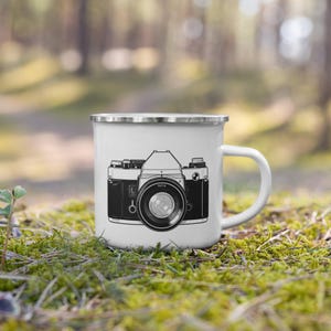 Può includere: Tazza smaltata bianca con un'illustrazione in bianco e nero di una fotocamera vintage. La tazza ha un bordo argentato e un manico bianco. Il design della fotocamera presenta un obiettivo, quadranti e altri dettagli.