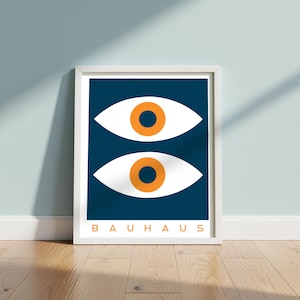 Könnte beinhalten: Ein gerahmter Kunstdruck mit minimalistischem Design. Er zeigt zwei weiße Augenformen mit orangefarbenen Iris auf einem dunkelblauen Hintergrund. Das Wort "BAUHAUS" ist unten in Orange gedruckt. Das Kunstwerk wird auf einem Holzboden ausgestellt.