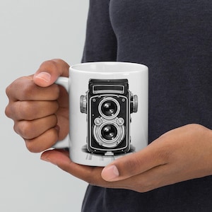 May include: White ceramic mug featuring a detailed black and white illustration of a vintage twin-lens reflex camera. The camera design includes intricate details of the lens, dials, and body. The mug has a standard handle.