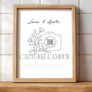 Peut inclure: Un cadre en bois clair contenant une impression en noir et blanc. L'impression représente un appareil photo avec un code QR, des fleurs, et les textes "Lucas & Agathe" et "CAPTUREZ L'AMOUR".