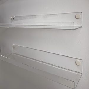 May include: Clear acrylic floating shelves fixed to a white wall. The shelves are rectangular with a front lip and are secured with white circular fixings. The design is minimalist and modern, suitable for various home decor styles.