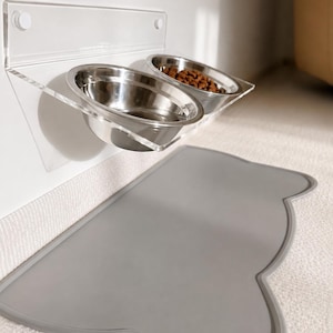 Puede incluir: Un comedero de pared de acrílico transparente con dos cuencos circulares de acero inoxidable, uno de ellos con comida seca marrón. Debajo del comedero hay una alfombrilla de silicona gris claro con un borde en forma de orejas de gato.