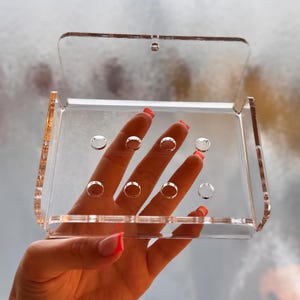 May include: A transparent acrylic box with a hinged lid. The box features several circular indentations on the front. The box is held up against a blurred background.