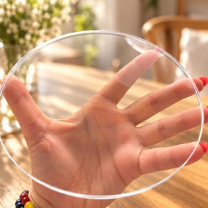 Pode incluir: Um disco circular de acrílico transparente com uma superfície lisa e clara e uma borda fina e arredondada. O disco é segurado na palma da mão, exibindo sua alta transparência e design plano e leve, permitindo ver através do material.