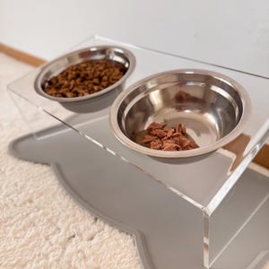 Könnte beinhalten: Ein klarer Acryl-Futternapf für Haustiere mit zwei Edelstahlnäpfen, einer gefüllt mit Trockenfutter und der andere mit Nassfutter. Der Futterautomat steht auf einer grauen, knochenförmigen Matte, um Verschüttungen aufzufangen.