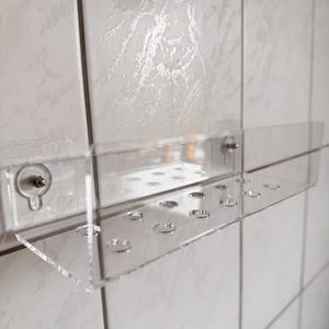 May include: Clear acrylic shelf with multiple circular holes, mounted on a white tiled wall. The shelf has a rectangular shape and is held in place by clear, round fasteners. The design is minimalist and functional.