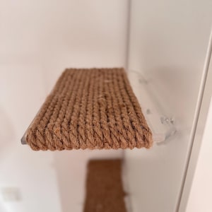 May include: A rectangular wall-mounted shelf featuring a thick, textured brown rope surface wrapped tightly around a base. The shelf is attached to a white wall with clear, transparent brackets, providing a sturdy platform for climbing or resting.