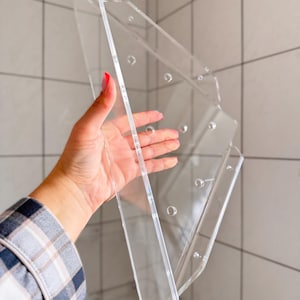 Könnte beinhalten: Ein klarer Acryl-Displayständer mit mehreren Paneelen und Bohrlöchern. Der Ständer wird von einer Hand gehalten, die seine Transparenz und sein Design zeigt. Der Ständer ist für die Aufnahme von Gegenständen konzipiert.