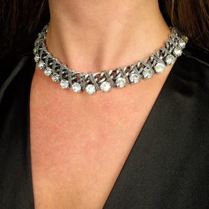 Puede incluir: Un collar gargantilla plateado con piedras preciosas redondas y transparentes. El collar presenta un diseño similar a una cadena con filas alternas de piedras preciosas y eslabones metálicos. La joya se usa sobre un fondo negro.