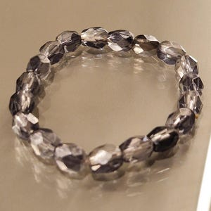 Puede incluir: Un brazalete de cuentas claras y facetadas. Las cuentas de forma ovalada están ensartadas para formar un círculo. El brazalete es de color gris claro y refleja la luz.